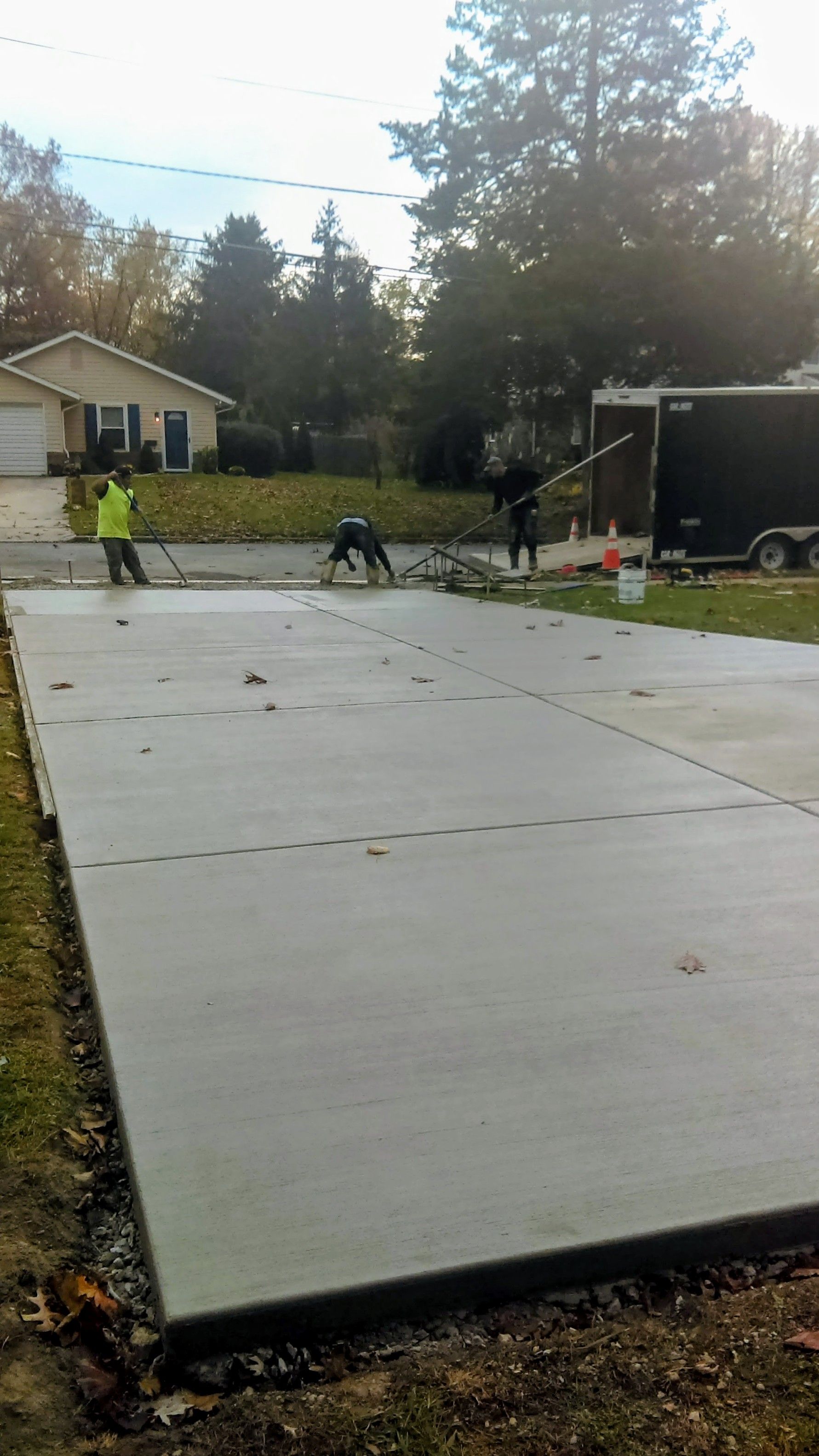 Construction workers smoothing wet concrete driveway, next to a house and trailer.