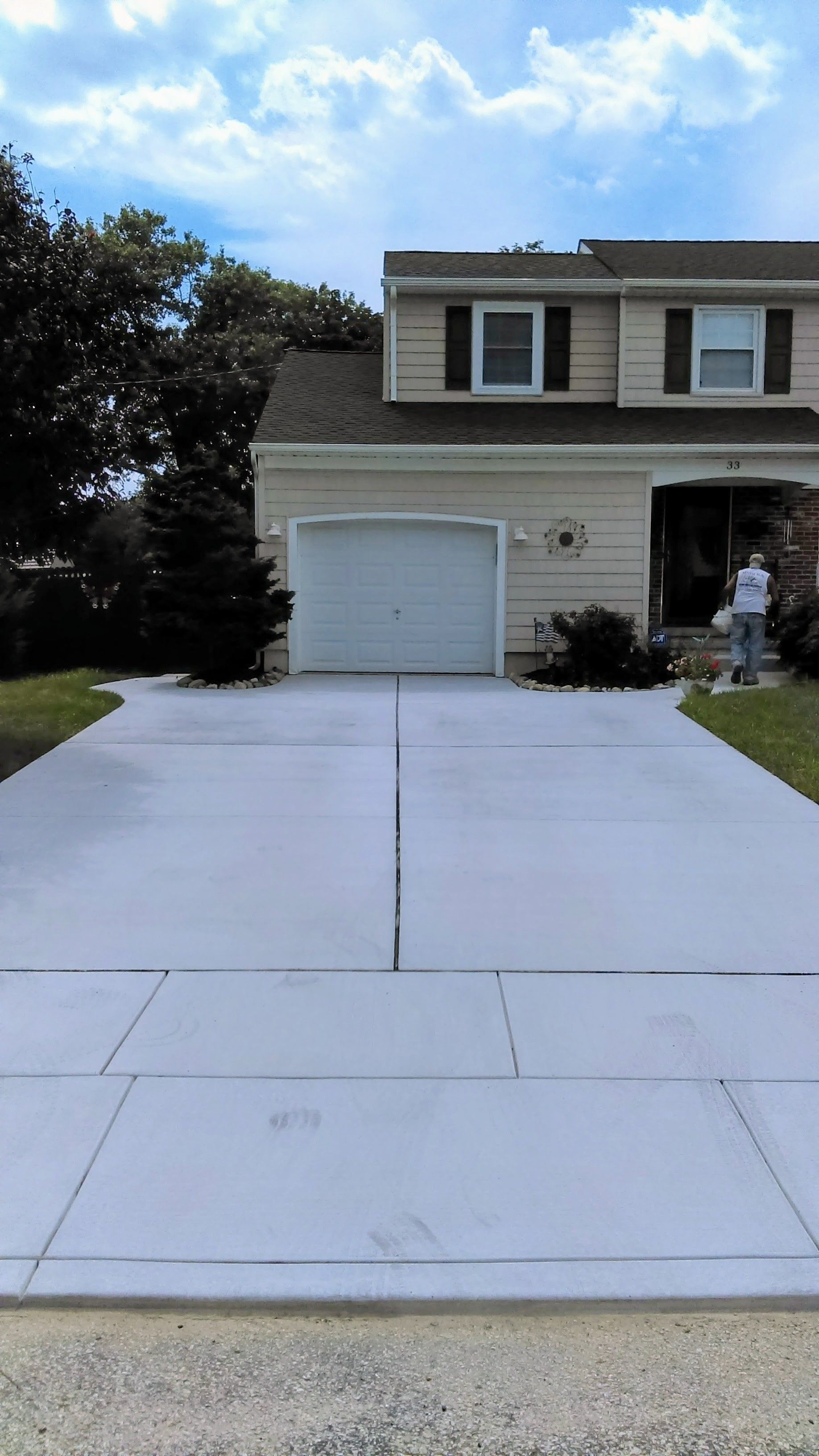 Newly poured concrete driveway in front of a two-story beige house with a garage; person in the doorway.