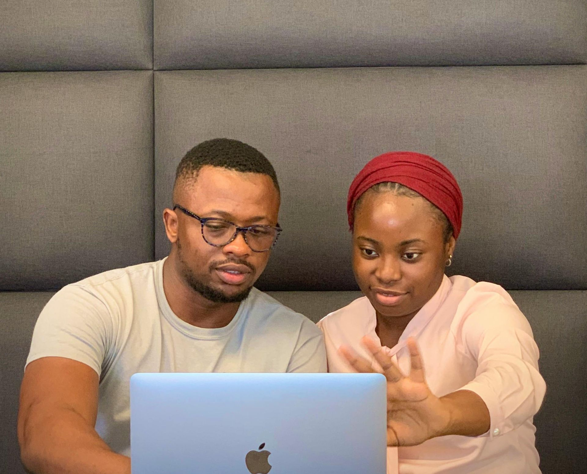 Man and woman looking at laptop, woman gesturing. They are indoors in front of a gray wall.