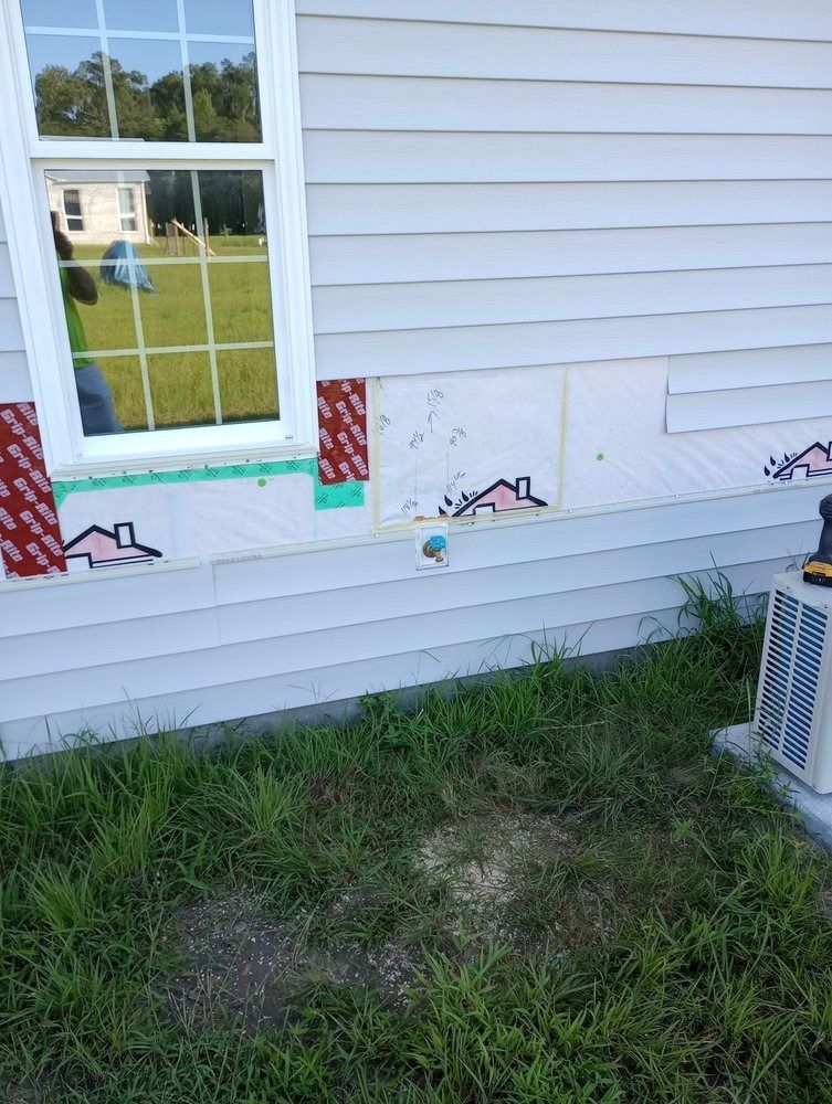 A house exterior with missing vinyl siding, revealing white insulation board, near a window and an outdoor AC unit.