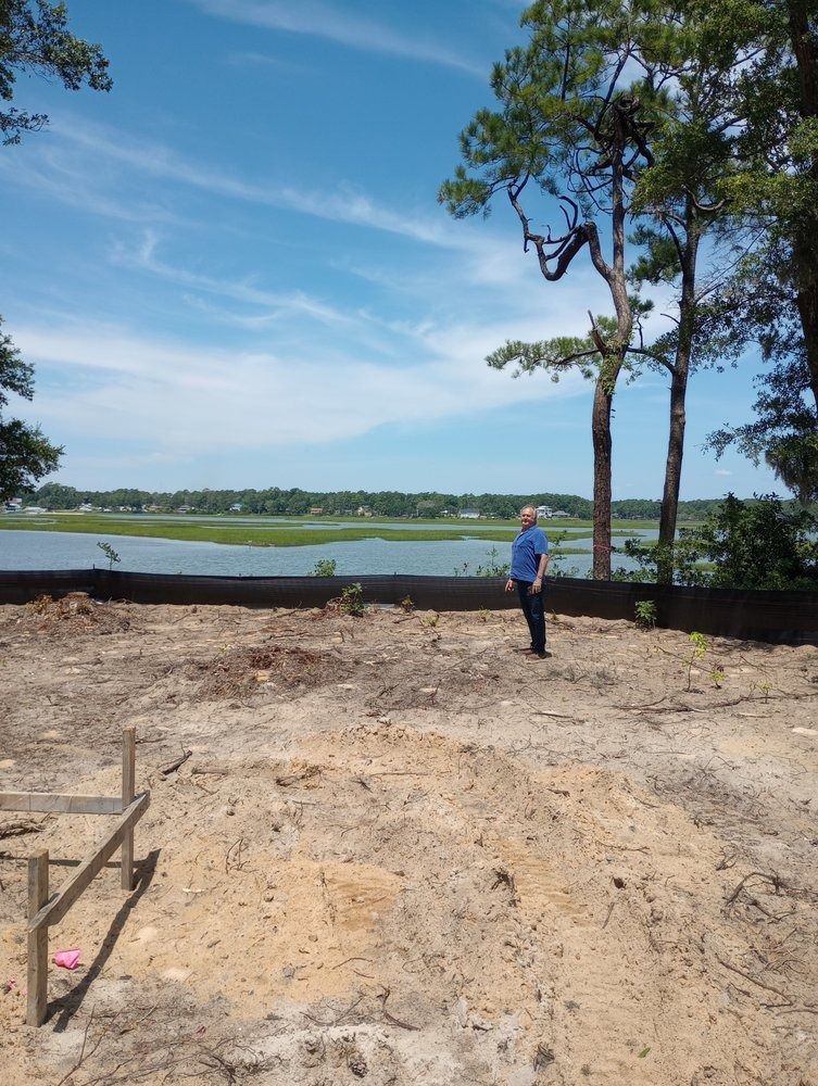 A person stands on a sandy construction site overlooking a grassy marshland under a bright blue sky.