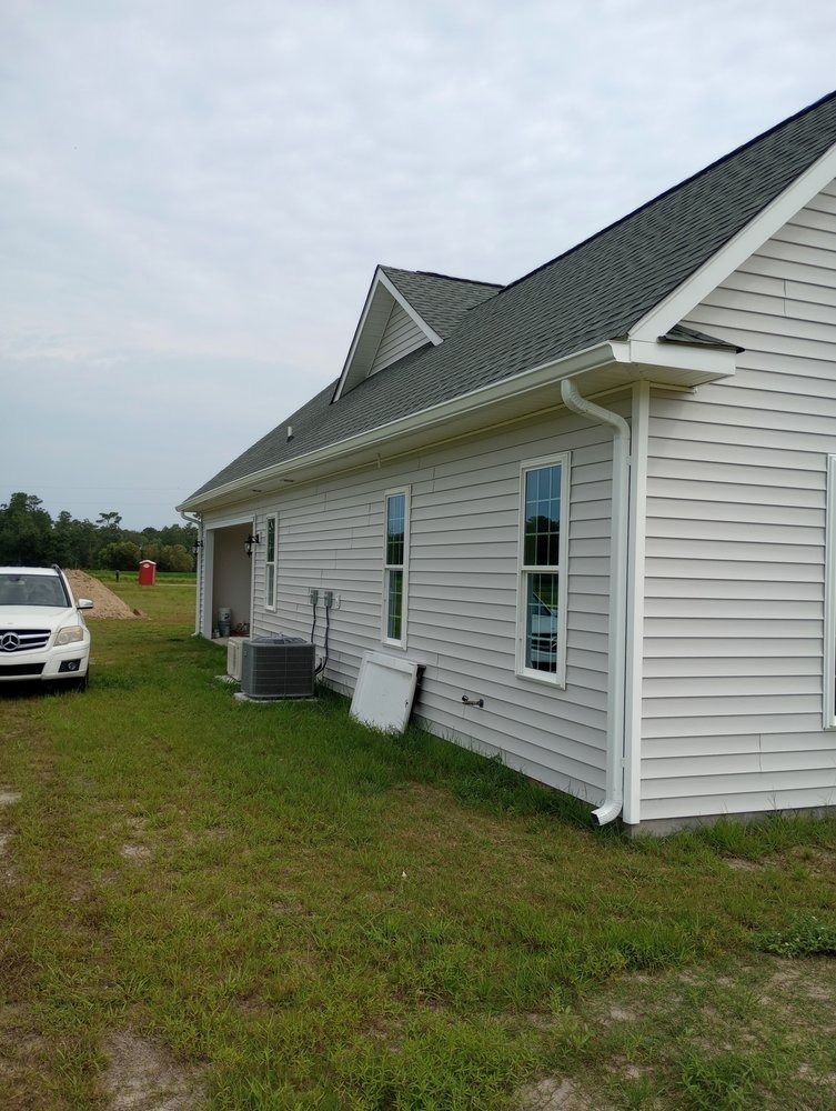 A light-colored house with a dark roof and white siding, viewed from the side on a grassy lot, with a white car nearby.