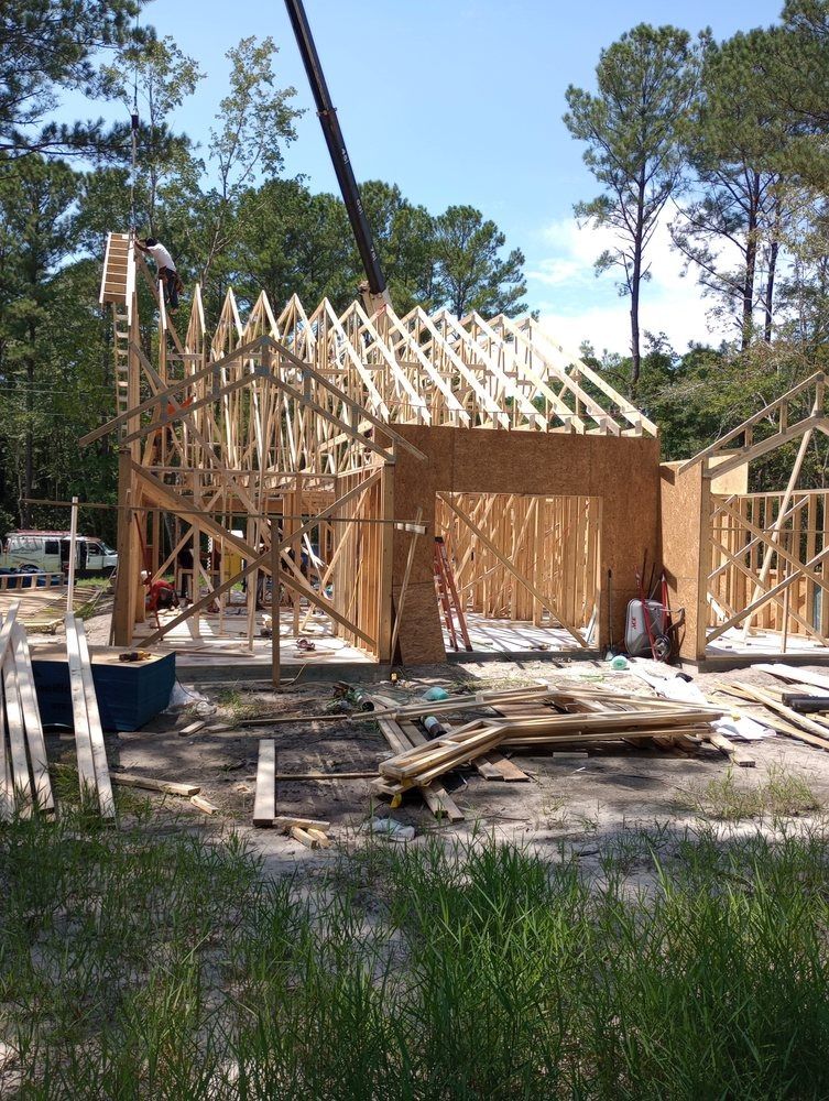 Wooden framing of a new house under construction in a wooded area, with a construction worker visible on the roof.