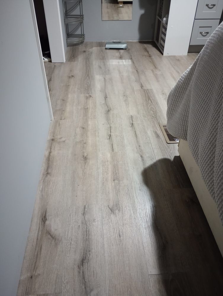 A perspective view of a light wood-look laminate floor in a bedroom, with a bed and furniture visible on the sides.