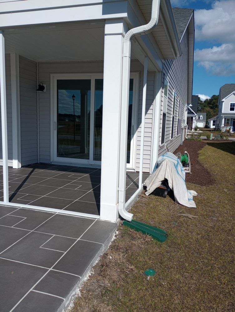 A covered patio with dark tiles, a white sliding door, and a white downspout leading to a green drain extension on the lawn.