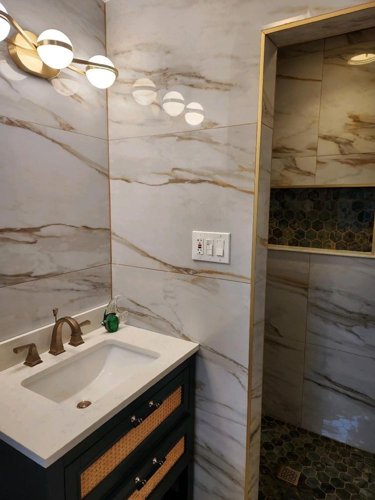 A bathroom vanity with a white countertop and gold faucet sits next to a tiled shower stall with gold trim.