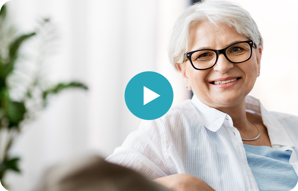 An elderly woman wearing glasses is smiling while sitting on a couch.
