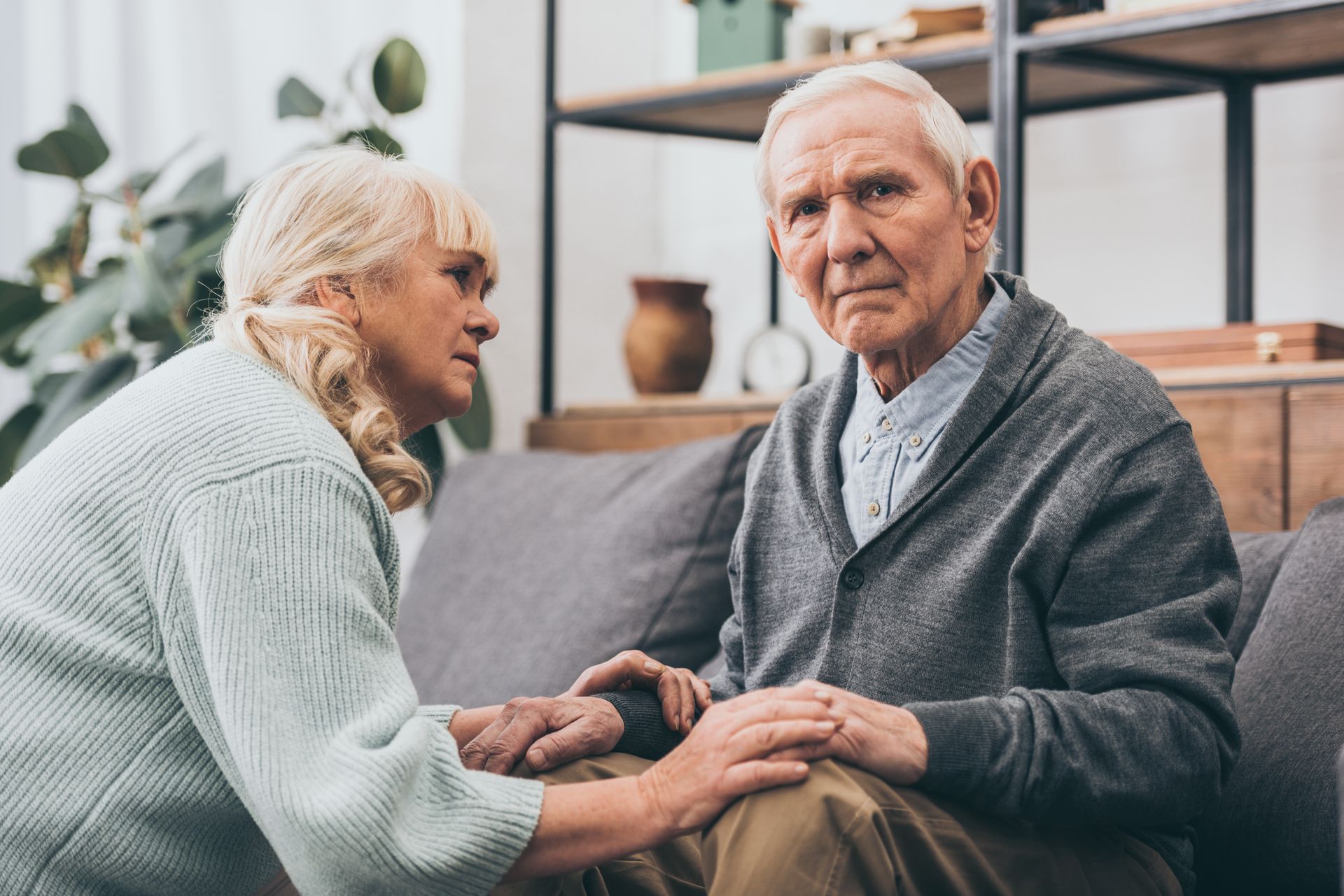 Woman comforts older person on couch, holding hands; concerned expression, indoor setting.
