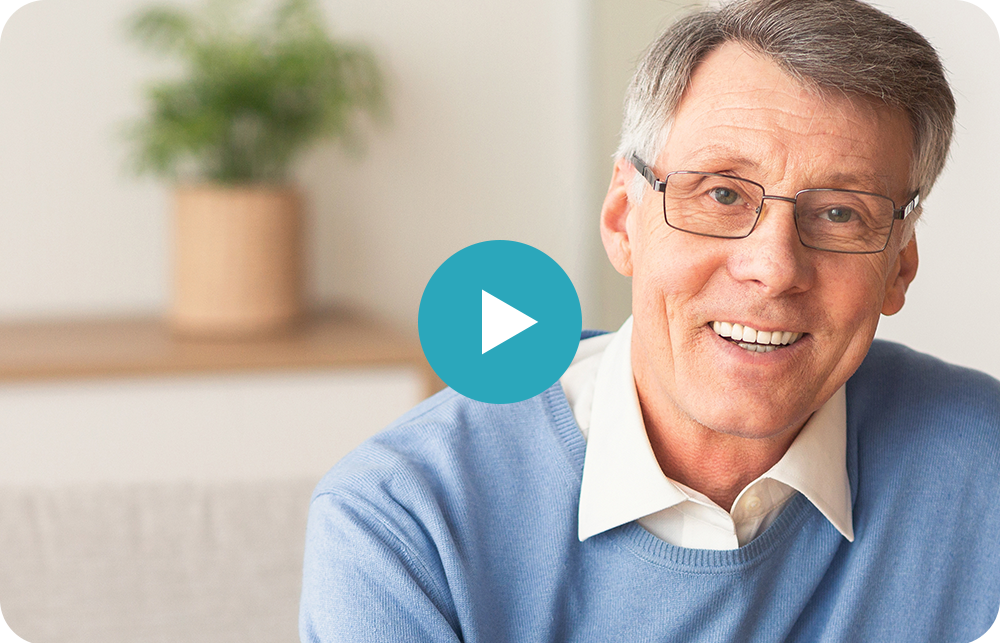 Smiling man in blue sweater with a play button overlay, seated indoors with a blurred plant in the background
