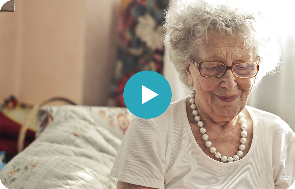 An elderly woman wearing glasses and a pearl necklace is sitting on a bed.