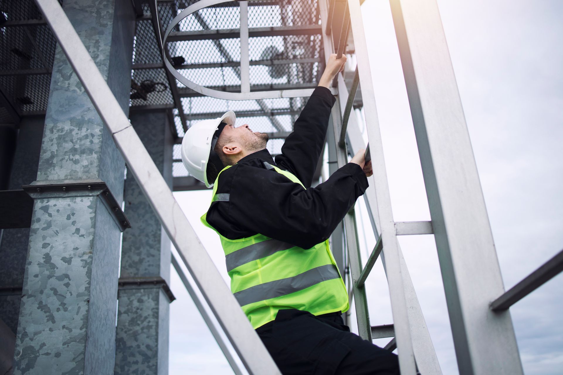 Construction worker in a hard hat and safety vest climbing a metal ladder on an industrial structure.