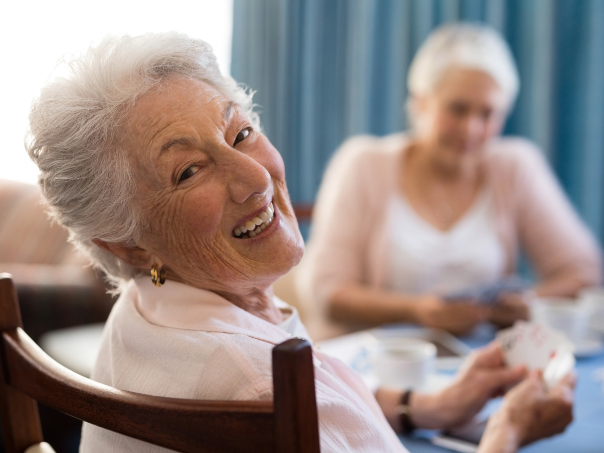 Smiling elderly woman in a chair, with another woman blurred in the background at a table representing support for Aged Care Faciltiies