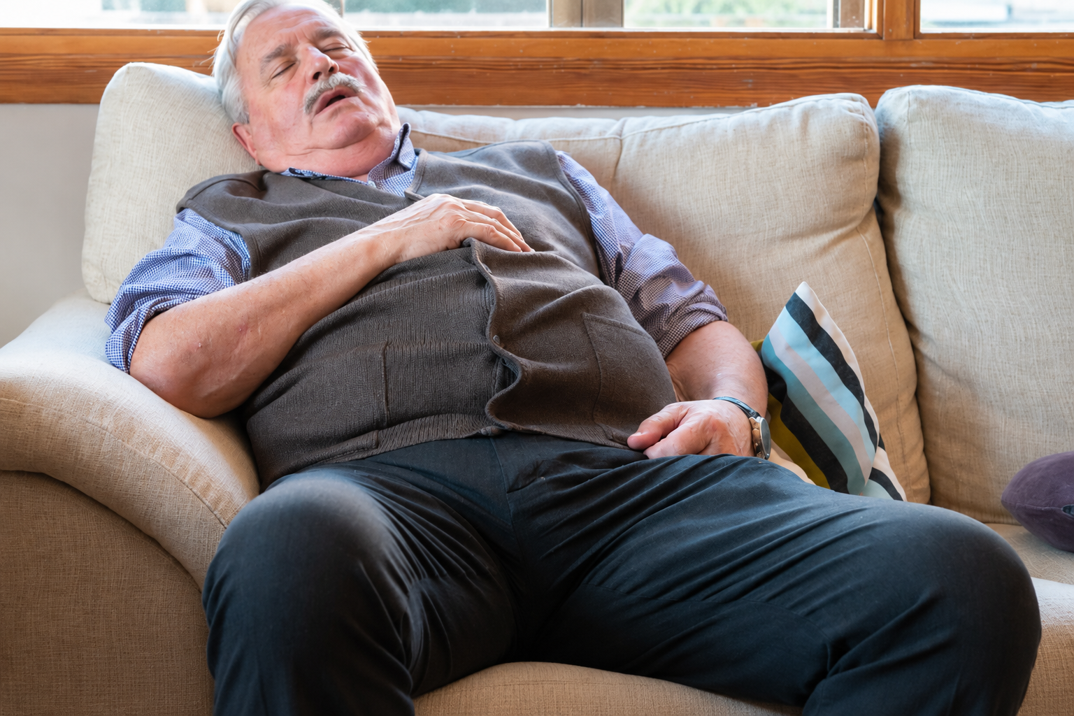 Man lying asleep on a beige sofa with a gray pillow, in a sunlit living room