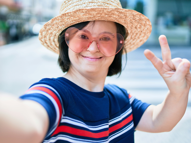 Person in a straw hat and pink glasses taking a selfie and making a peace sign outdoors representing support for NDIS participants