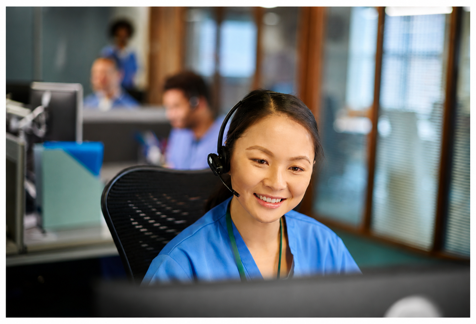 Customer support agent wearing a headset, smiling at a desk in an office with coworkers in the background