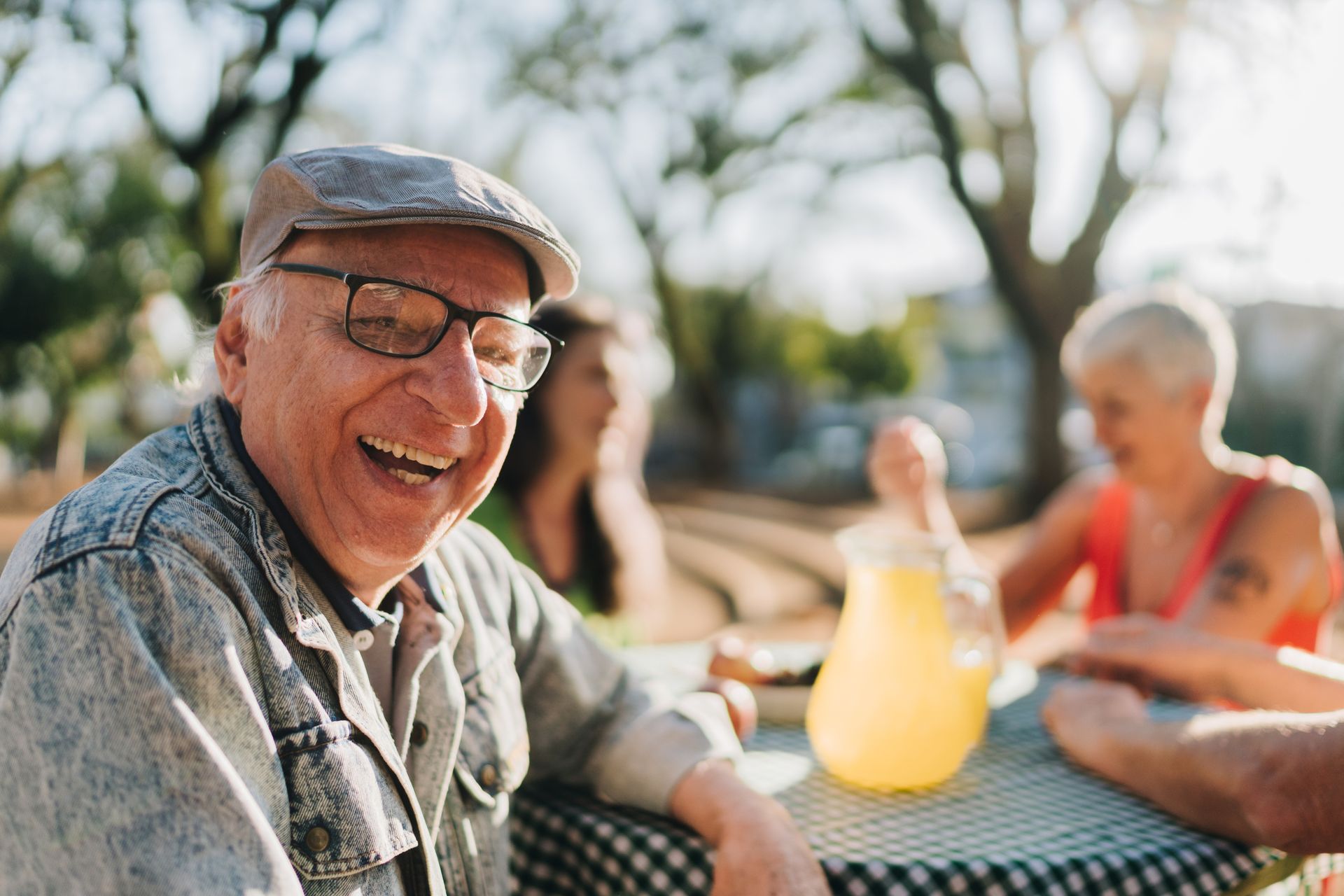 Smiling man in glasses and cap at an outdoor table with drinks, chatting with others in sunlight representing senior support