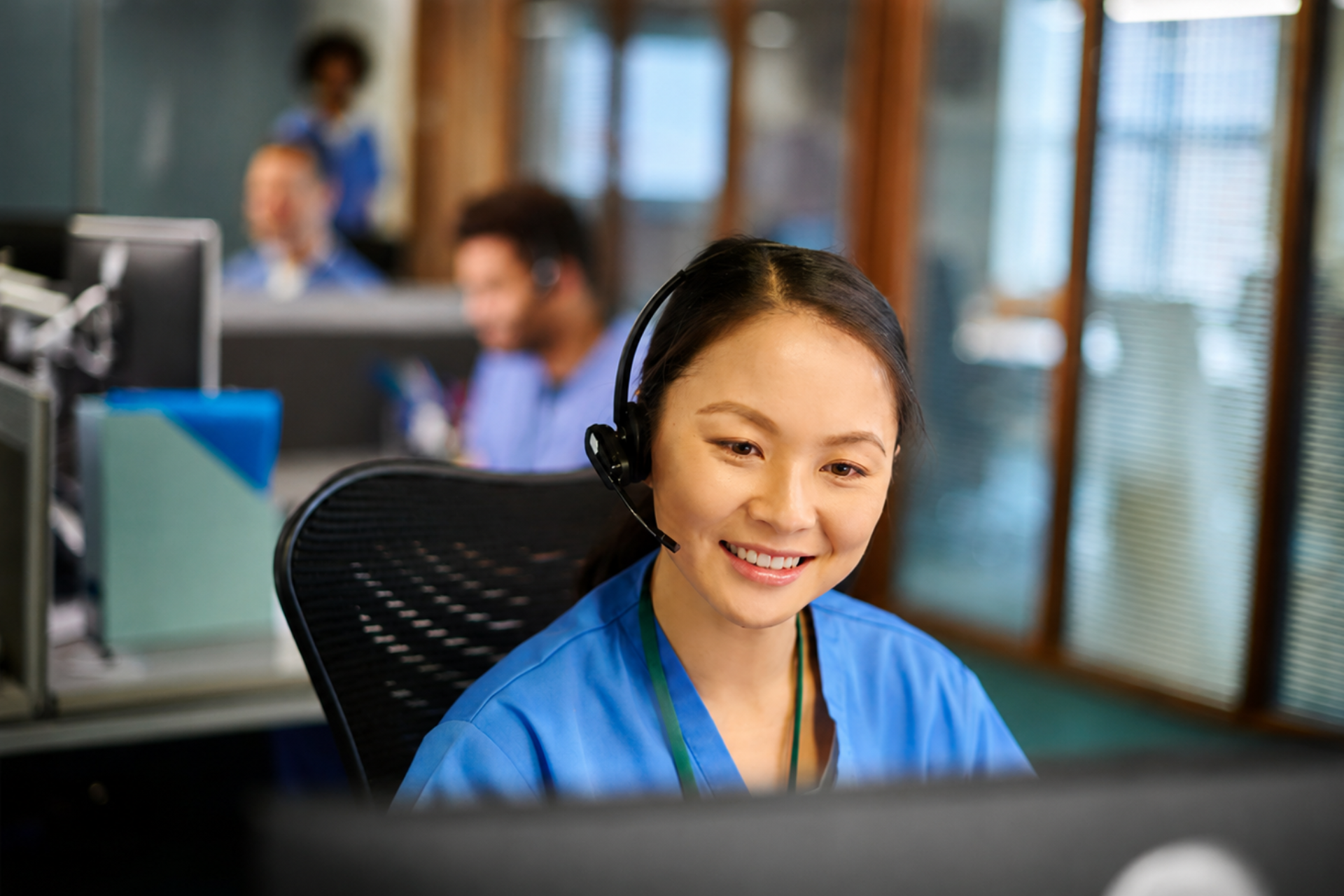 Customer support agent wearing a headset, smiling at a desk in an office with coworkers in the background