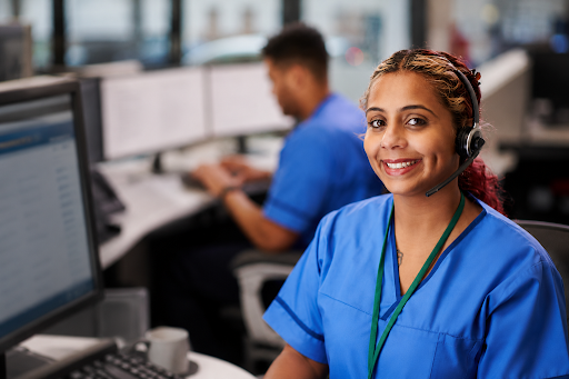Smiling nurse who is an emergency agent in blue scrubs wearing a headset in an office, with a coworker working behind her