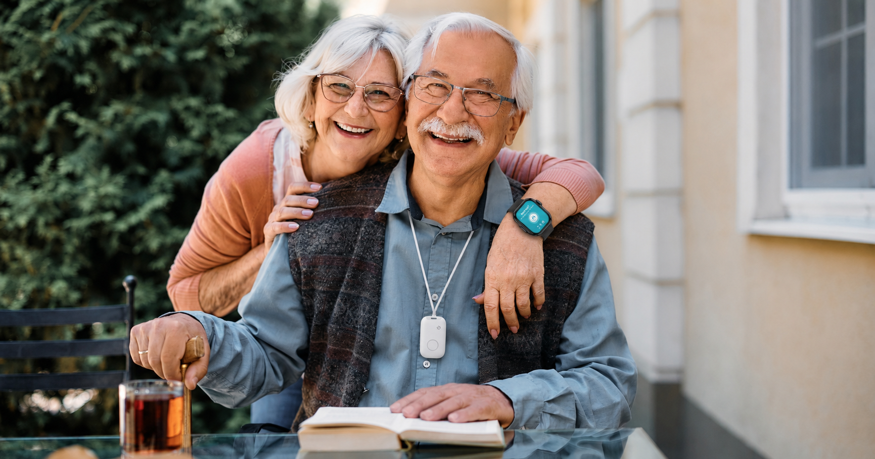 A smiling older lady wearing a smart watch, a personal alarm, and an smiling older man seated wearing a personal alarm around his neck, outdoors with a book and a drink on a patio