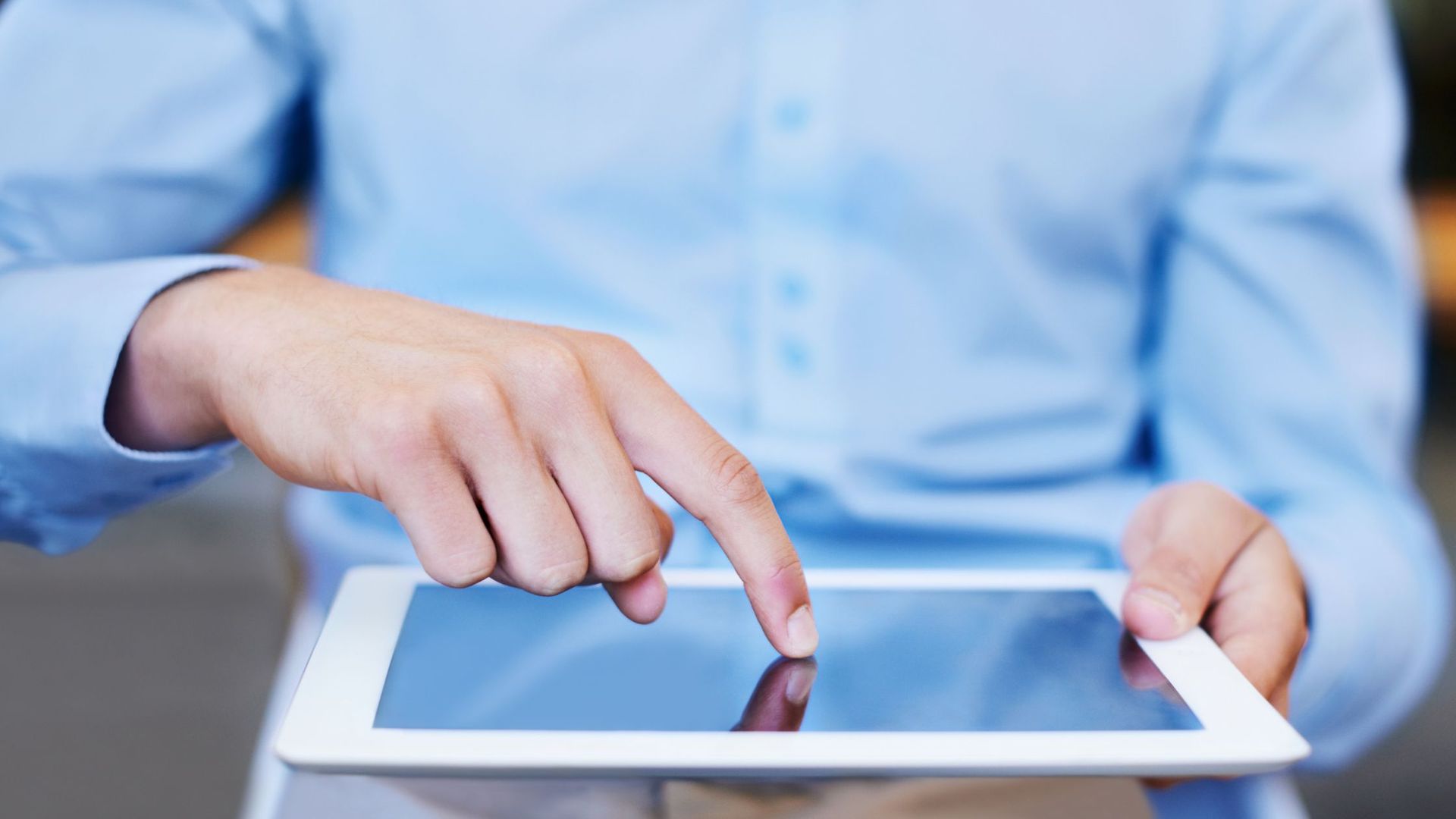 Person in blue shirt using a white tablet with index finger, close-up shot.