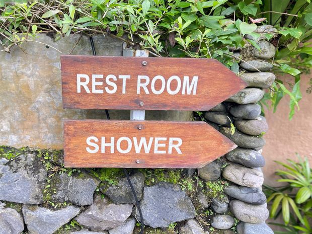 Wooden signs reading “REST ROOM” and “SHOWER” beside a stone wall and greenery