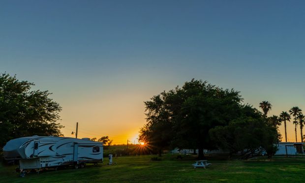 Sunset over a grassy campsite with a white camper trailer, trees, and a glowing horizon