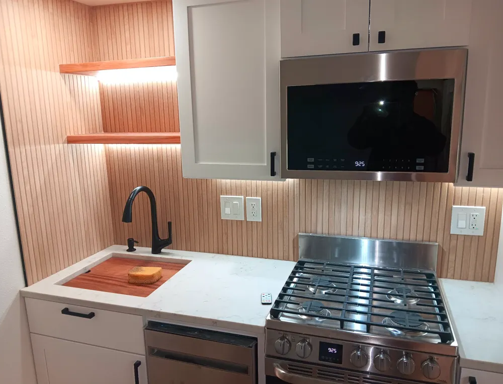Modern kitchen corner with black sink, white cabinets, wood backsplash, stainless stove, and microwave.