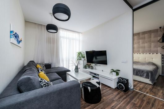 Modern living room with gray sectional sofa, wall-mounted TV, and a bedroom visible through an open doorway