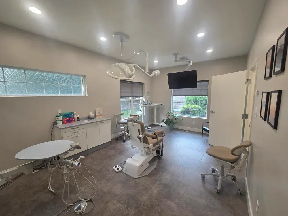 Medical exam room with exam chair, round table, stools, cabinets, and wall-mounted monitor.