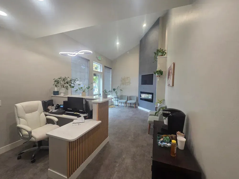 Modern office reception with desk, chairs, gray walls, and a wall-mounted fireplace leading to a bright back area