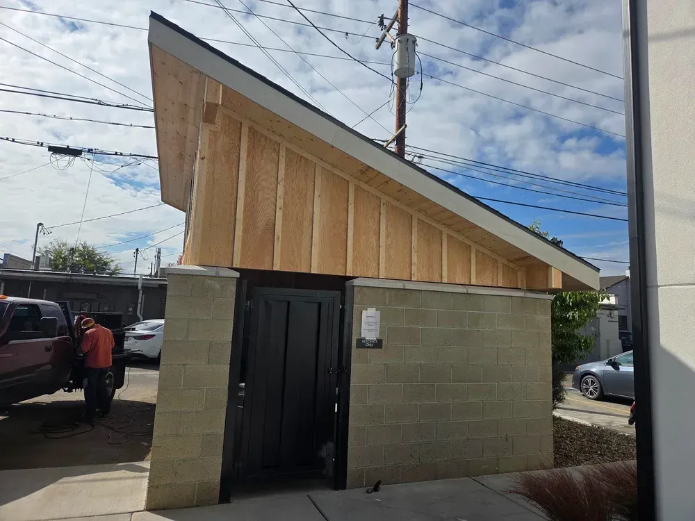 Small block building with a sloped wooden roof and black door beside a driveway under power lines.