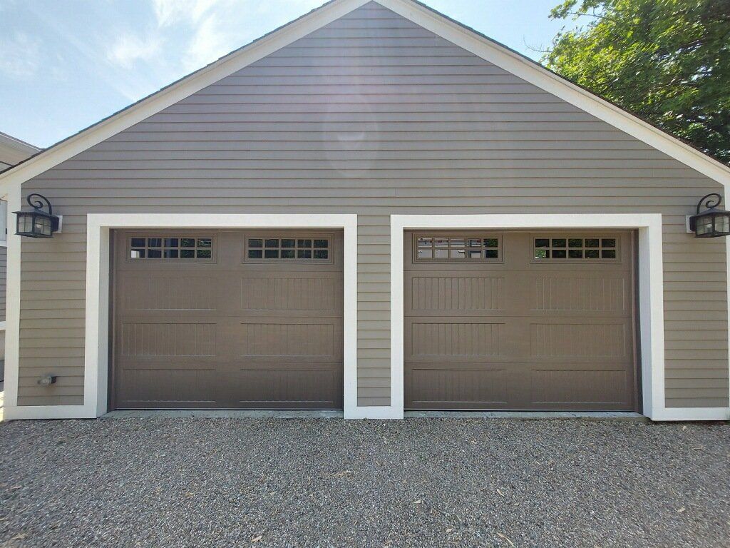 Two-car garage with brown doors, stone driveway, and gray siding. Outdoor lights mounted on the building.
