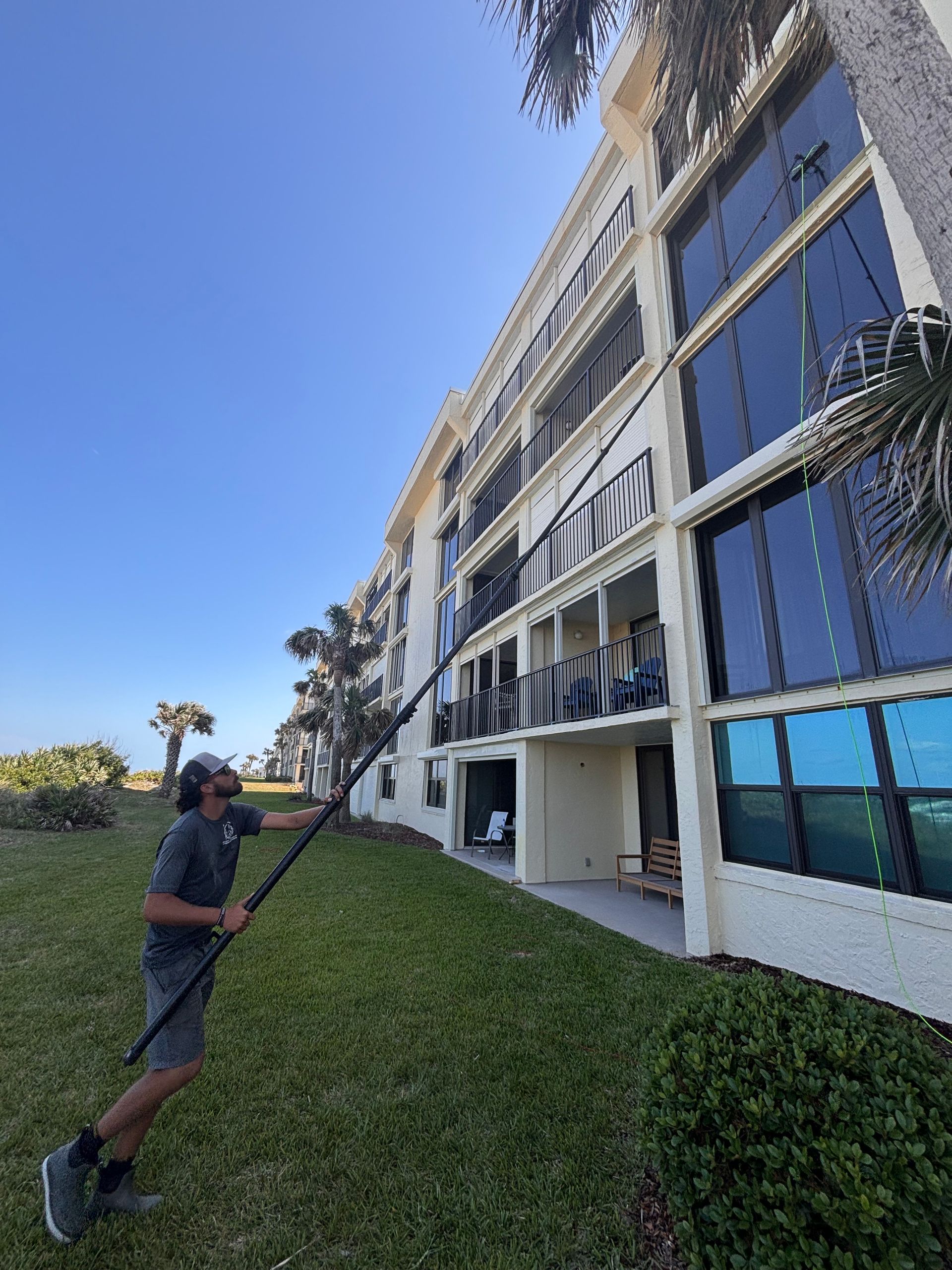 Man washing building windows with long pole, standing on grass. Sunny day.