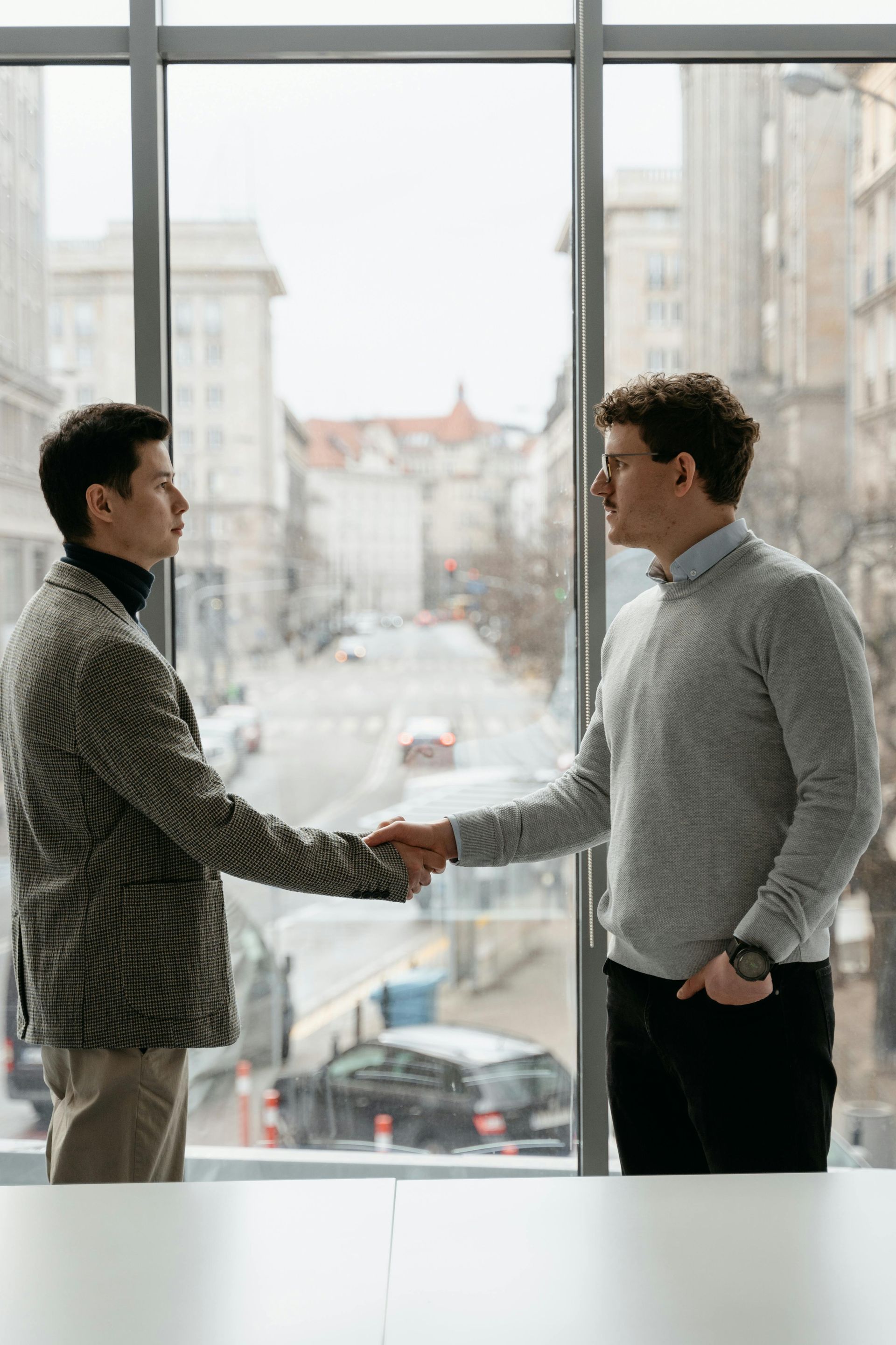 Two men shaking hands in front of a large window in a bright office.