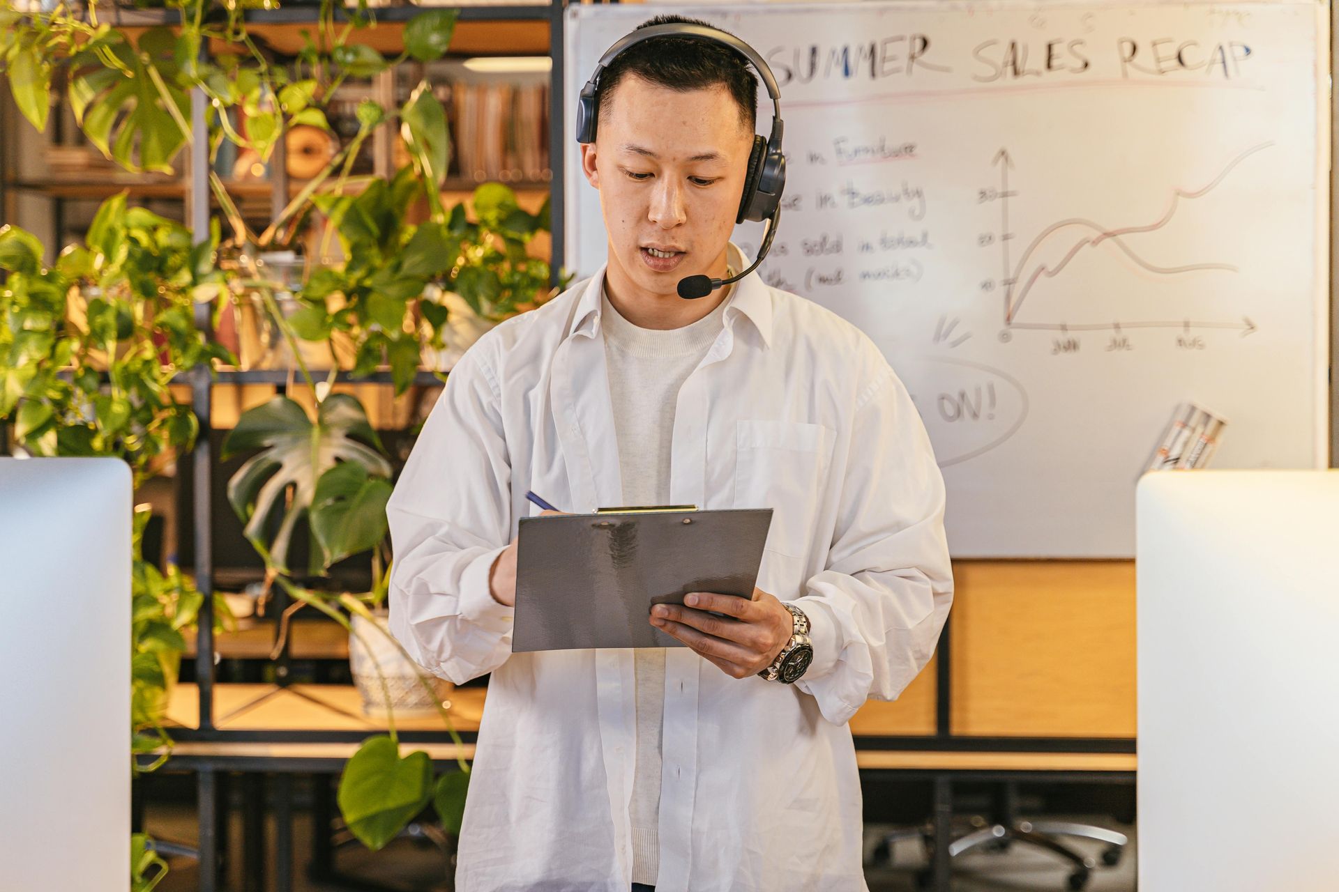Man in white shirt and headset reading from a tablet in a bright office with plants and a whiteboard.