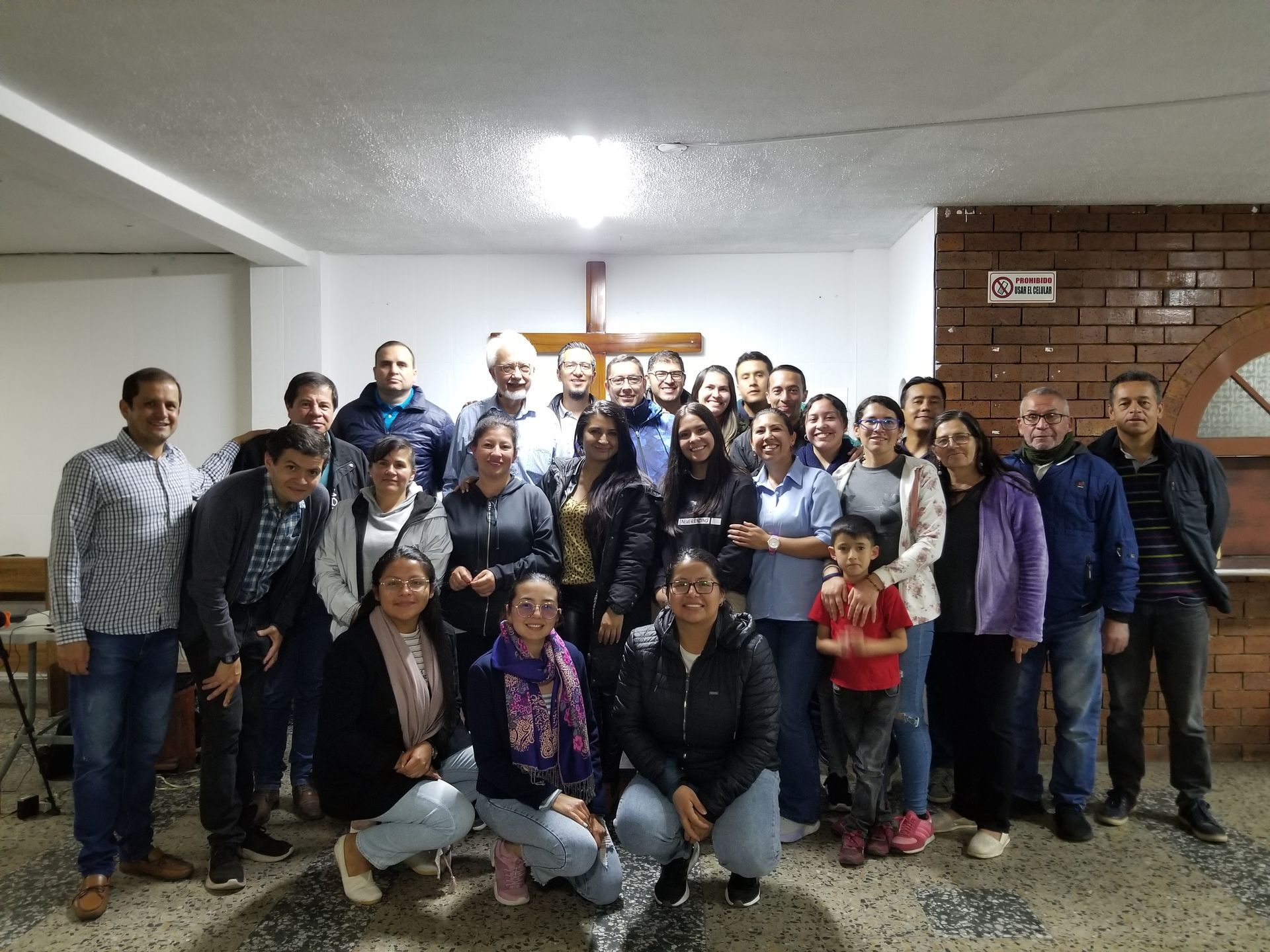 Group of diverse people gathered indoors; a cross hangs on the wall.