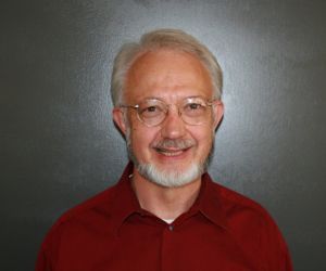 Man with gray beard and glasses smiling, wearing a red shirt, against a dark gray background.