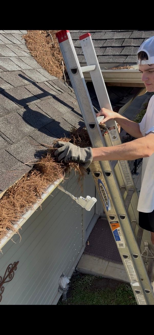 Worker on a ladder cleaning leaves from a rooftop gutter beside a house