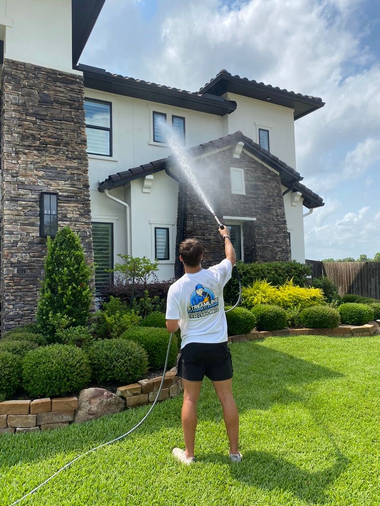 Person spraying a two-story house with a hose in a sunny front yard