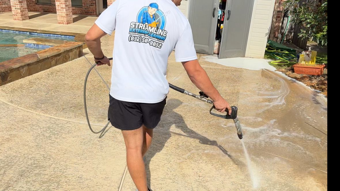 Worker pressure-washing a wet concrete driveway near a house and potted plants
