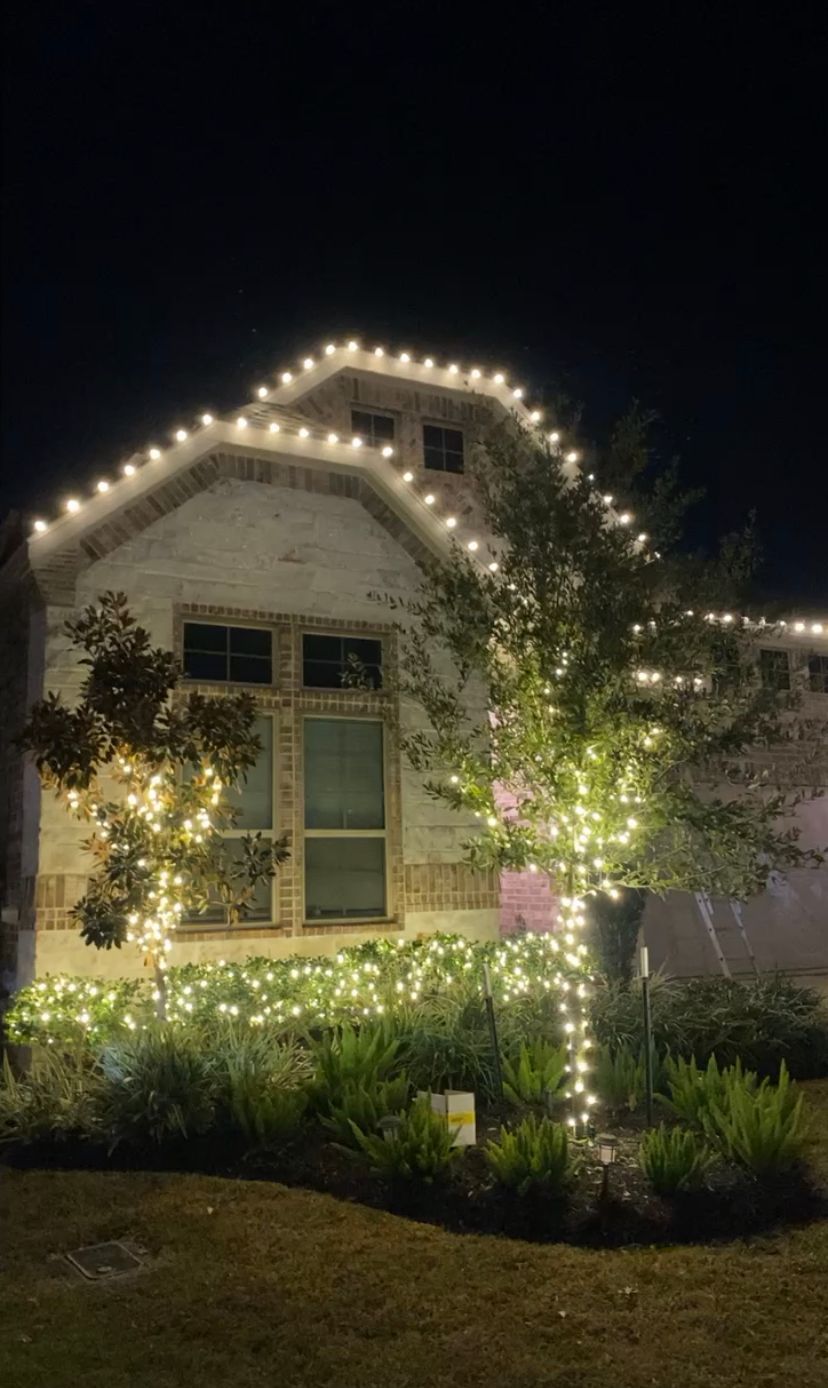 House at night outlined with warm white string lights and lit trees and shrubs in the front yard