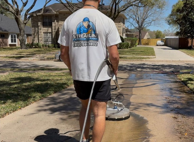 Worker pressure washing a driveway on a sunny suburban street, seen from behind