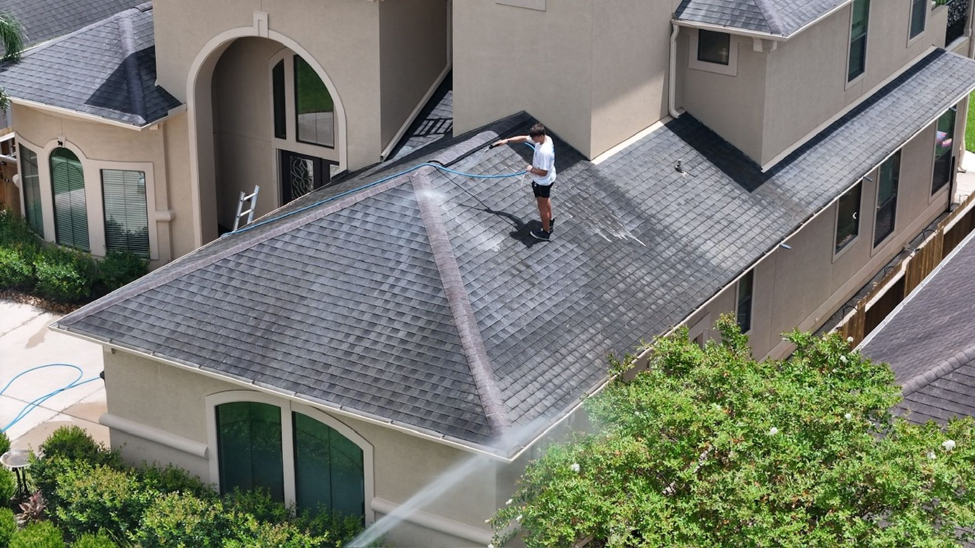 Worker repairing shingles on a gray roof of a beige house with an arched entryway.