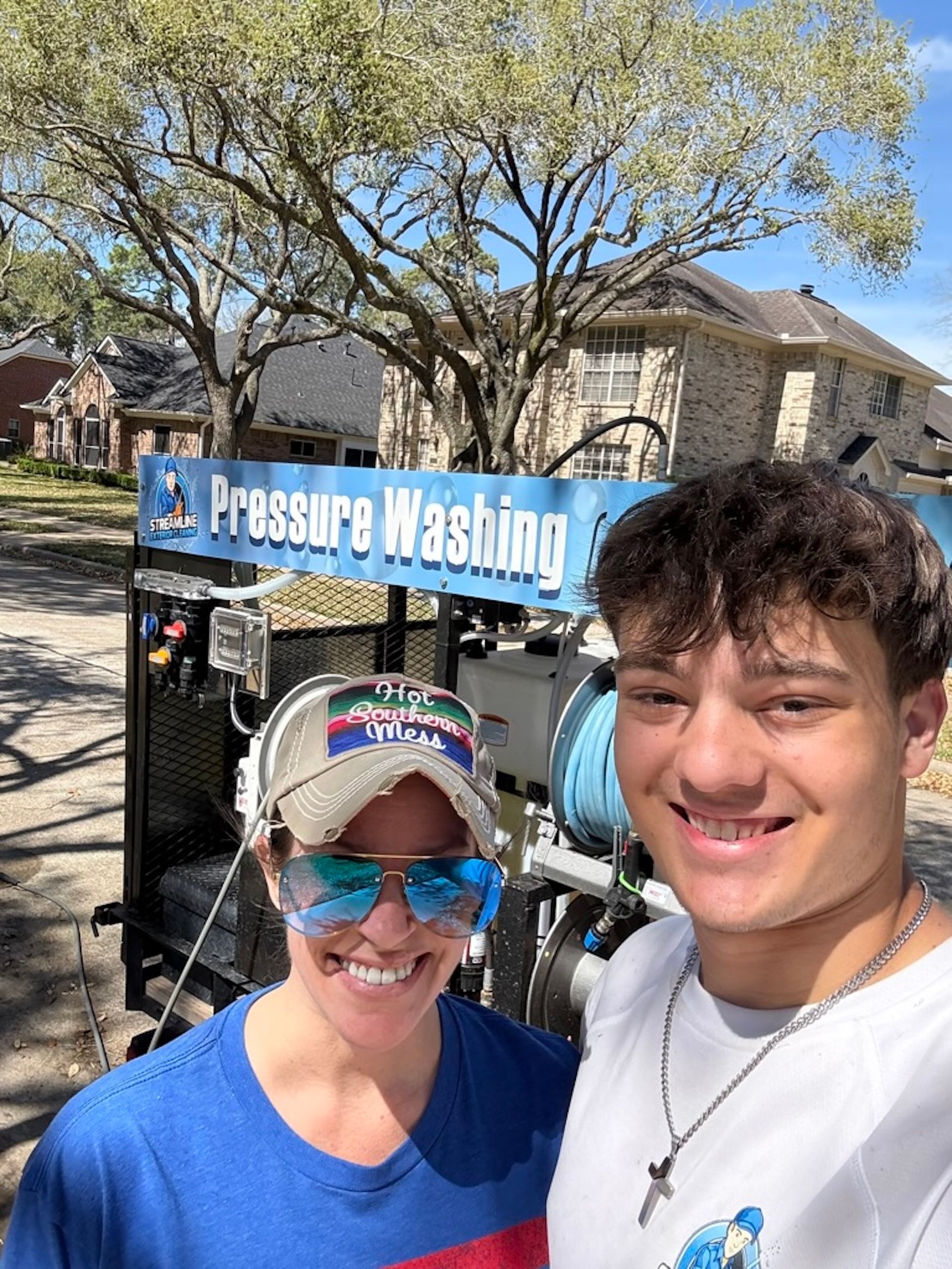Two smiling people posing in front of a pressure washing sign and equipment outdoors.