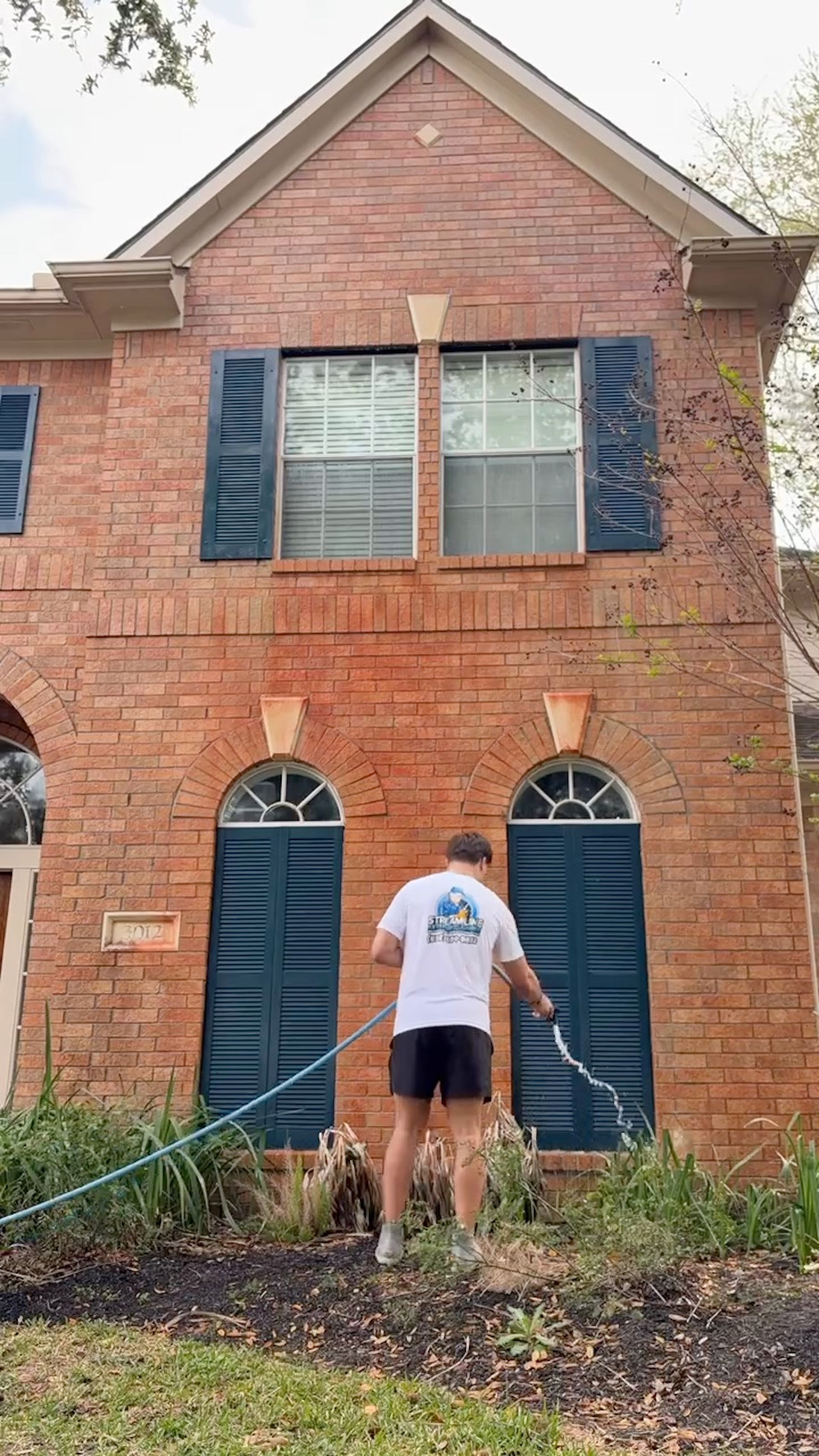Person standing in front of a brick house with blue shutters and double front doors, holding a hose.