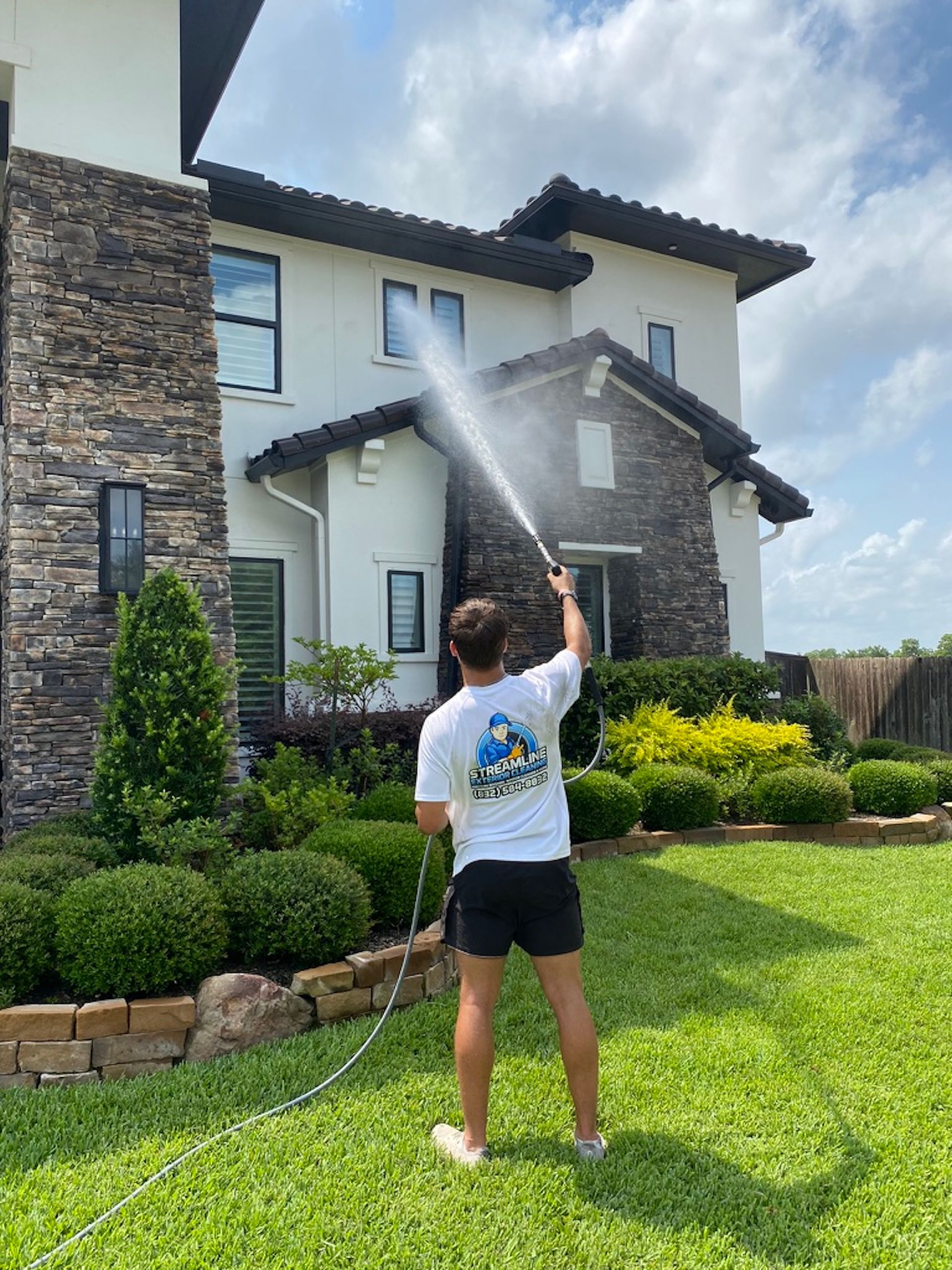 Person spraying water on a two-story house with a stone facade and green lawn