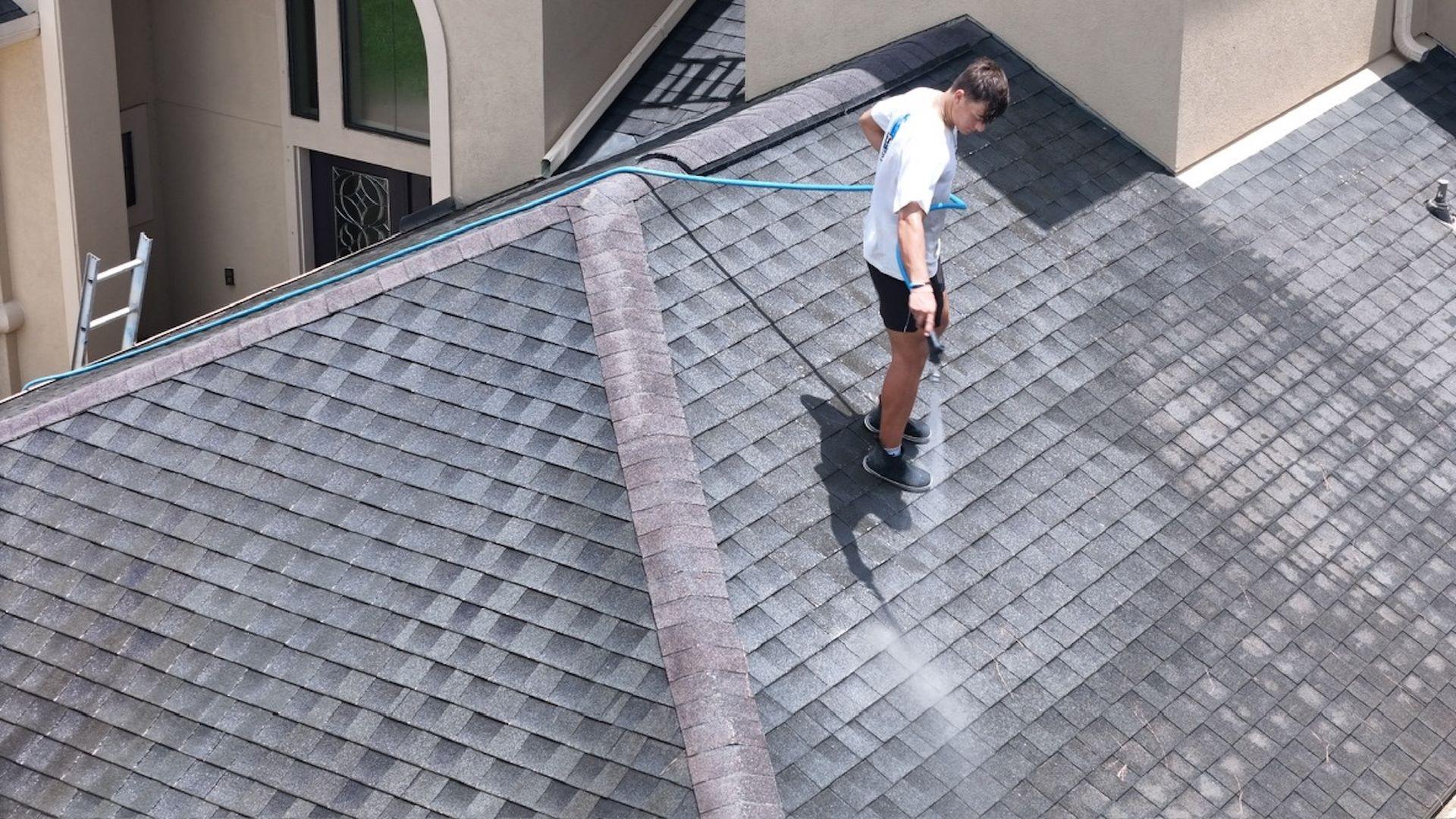Person standing on a gray shingled roof near a ridge line, looking downward.