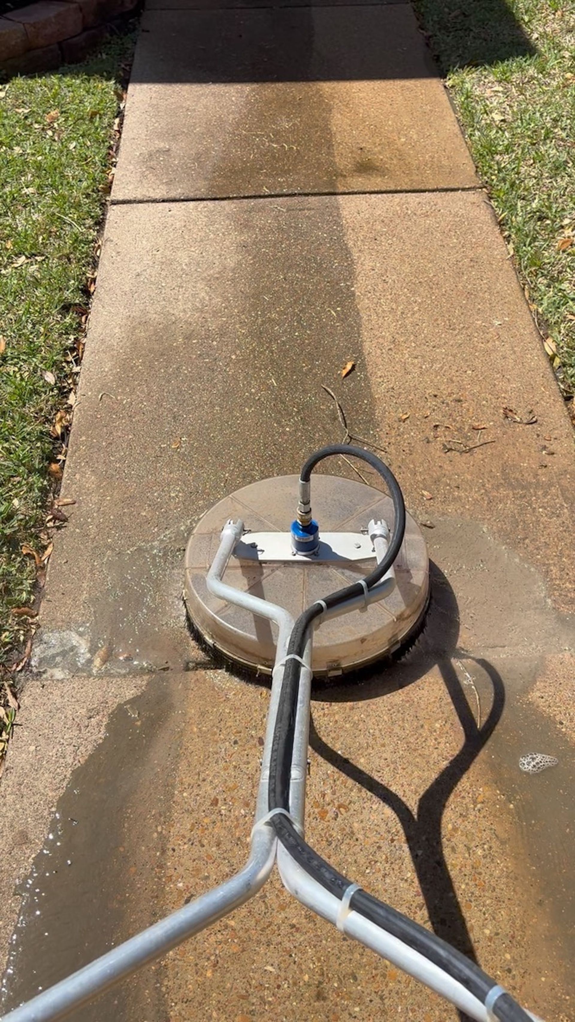 Pressure washer cleaning a concrete sidewalk, leaving a wet streak on the pavement.