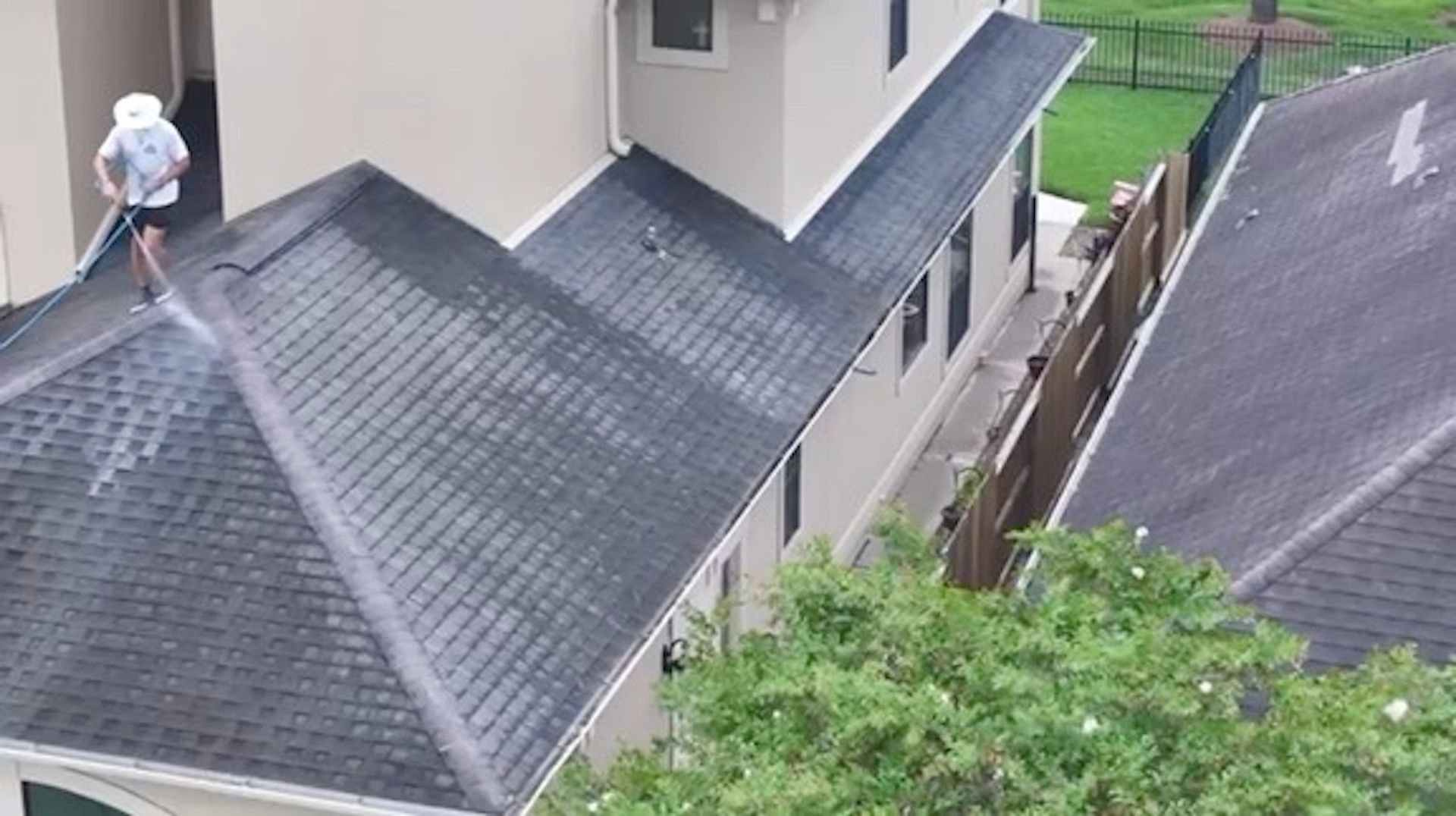 Worker on a steep gray roof using a safety harness near a house and trees