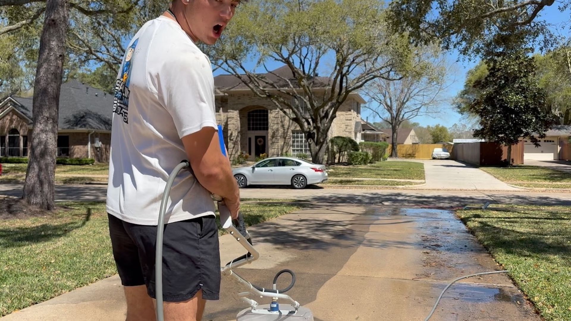 Person in white shirt and black shorts watering a driveway with a hose in a sunny suburban yard